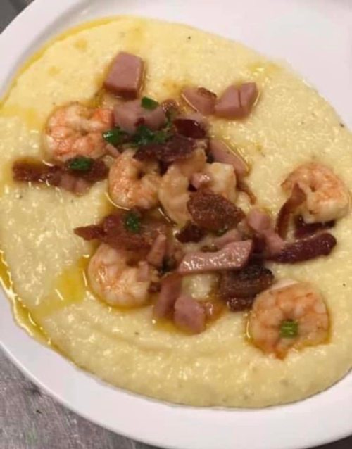A plate of creamy grits topped with shrimp, bacon, and diced ham, drizzled with olive oil.