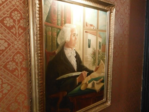 A framed portrait of a man with white hair, seated at a desk, surrounded by books and a window view.