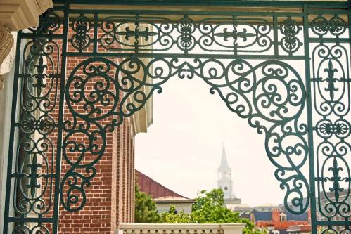 Ornate wrought iron gate framing a view of a church steeple and buildings in the background under a clear sky.