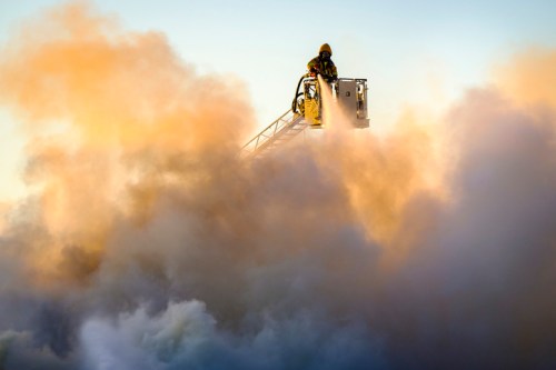 A firefighter on a ladder sprays water into thick smoke during a fire response, with a hazy, colorful sky in the background.