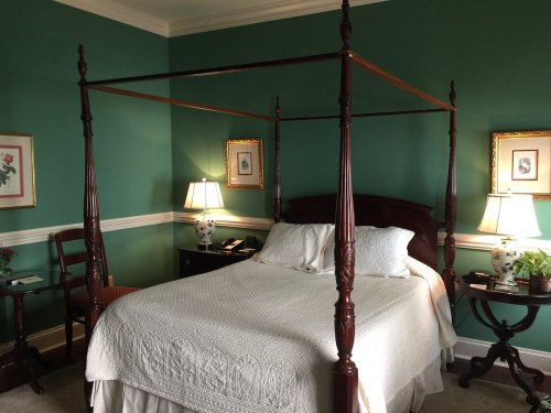 A cozy bedroom with a four-poster bed, green walls, two lamps, and decorative artwork.