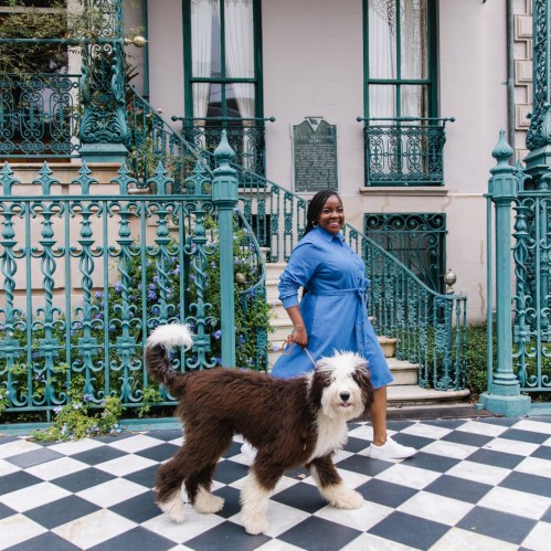 A woman in a blue dress stands beside a fluffy dog on a checkered patio in front of a decorative building.