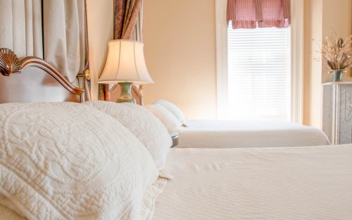 A cozy bedroom with two beds, soft white bedding, a lamp, and natural light streaming through a window.
