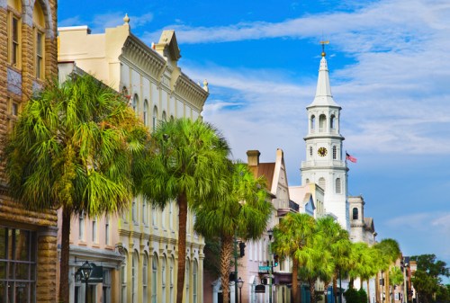 Historic buildings line a street with palm trees, featuring a tall church steeple against a blue sky.