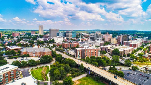 Aerial view of a city skyline with modern buildings, green spaces, and a highway under a blue sky with clouds.