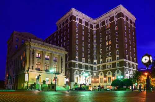 Historic buildings illuminated at night, featuring a clock and vibrant greenery in the foreground.