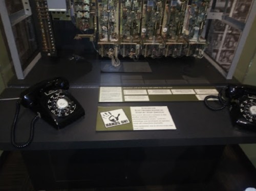 Two vintage black rotary phones on a display table, with a circuit board backdrop and an informational sign.