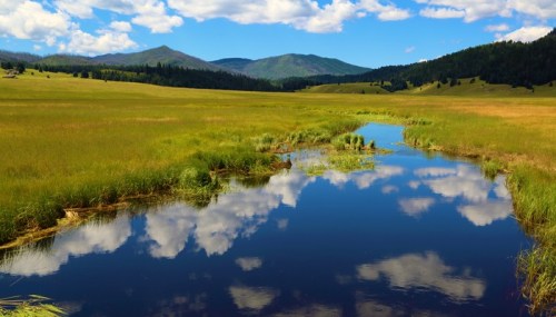 A serene landscape featuring a calm river reflecting clouds, surrounded by lush green fields and distant mountains.