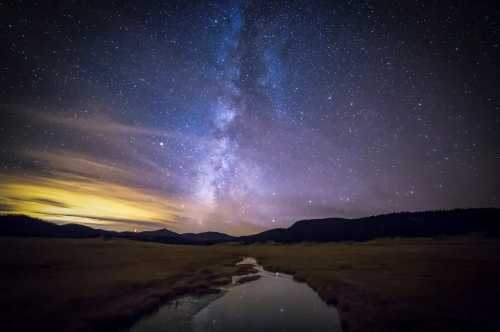 A serene night sky filled with stars and the Milky Way, reflecting in a calm stream amidst dark hills.