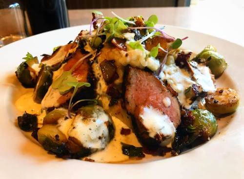 A plate of sliced meat topped with sauce, surrounded by Brussels sprouts and garnished with microgreens.
