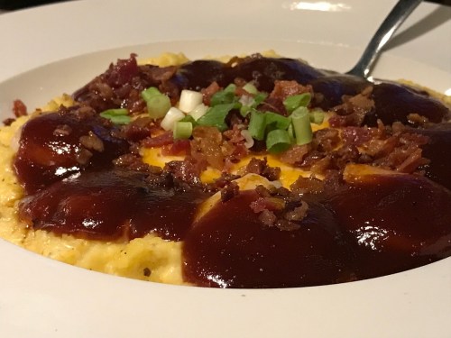 A bowl of creamy grits topped with barbecue sauce, bacon bits, and green onions, served with a spoon.