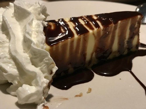 A slice of cheesecake drizzled with chocolate sauce, topped with a dollop of whipped cream on a plate.