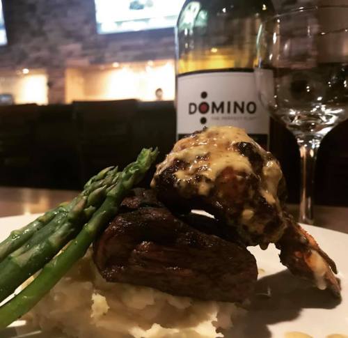 A plate of steak and shrimp topped with sauce, served with asparagus and mashed potatoes, alongside a glass of wine.