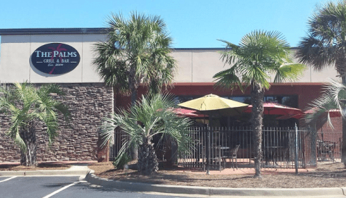 Exterior of The Palms Grill & Bar, featuring palm trees, outdoor seating with umbrellas, and a stone facade.