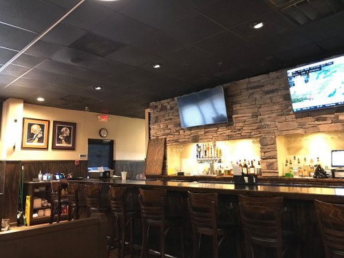 Interior of a bar featuring a stone wall, a long wooden counter, and a TV displaying a weather report.