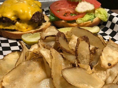 A cheeseburger with lettuce, tomato, and pickles, served with crispy potato chips on a checkered plate.