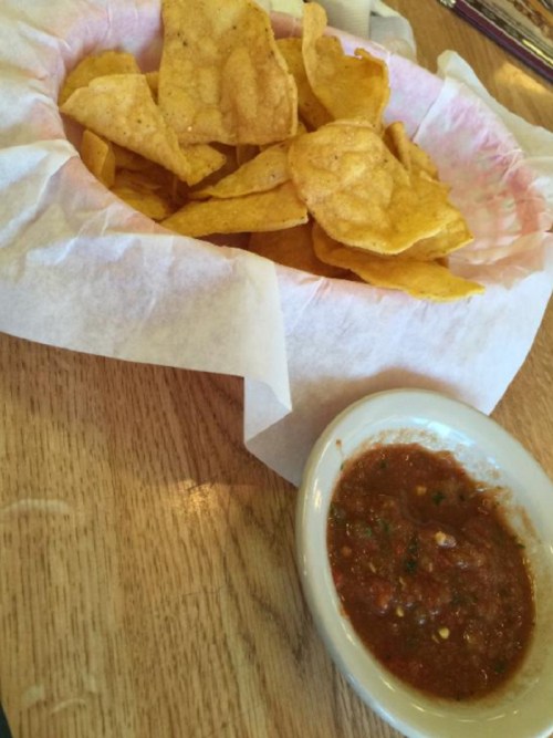 A basket of crispy tortilla chips next to a small bowl of salsa on a wooden table.