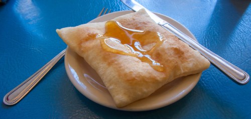 A golden-brown fried pastry drizzled with honey, served on a small white plate with a fork and knife.