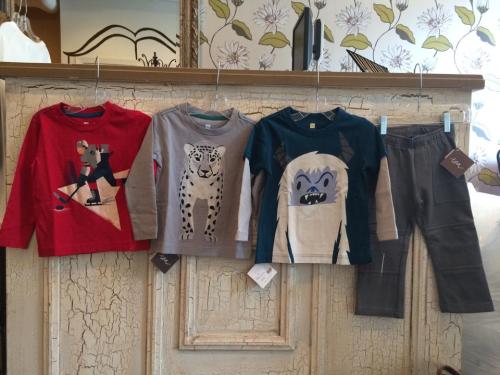 Four children's long-sleeve shirts featuring playful designs, hanging on a display with a pair of gray pants.