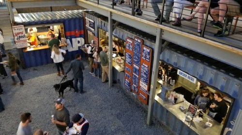 A lively outdoor food market with people at food stalls and a dog, surrounded by shipping containers and a balcony above.