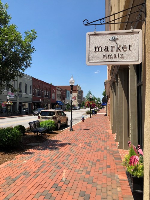 A sunny street view featuring a sign for "Market on Main," with brick sidewalks and shops lining the road.