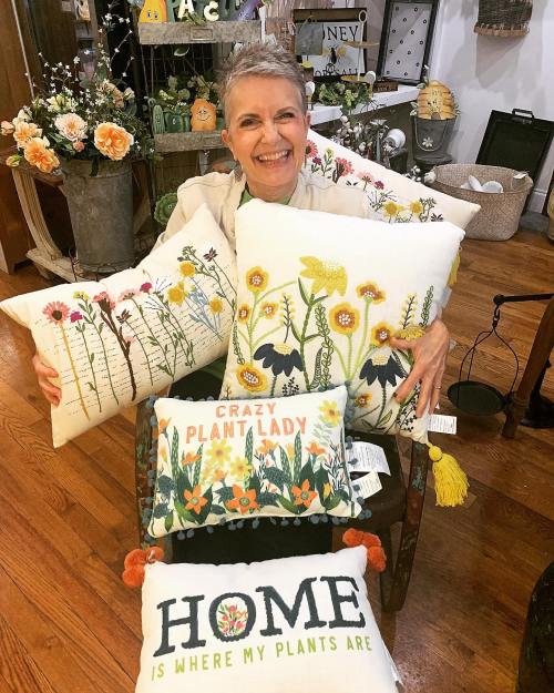 A smiling woman holds colorful decorative pillows featuring floral designs in a cozy shop setting.