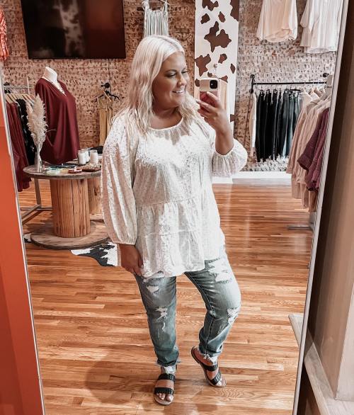 A woman in a white blouse and jeans smiles at her reflection in a store mirror, surrounded by clothing displays.