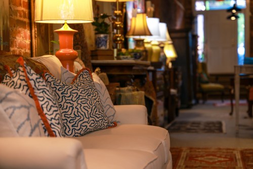 A cozy interior featuring a white sofa with patterned pillows, warm lighting, and a glimpse of a stylish decor shop.