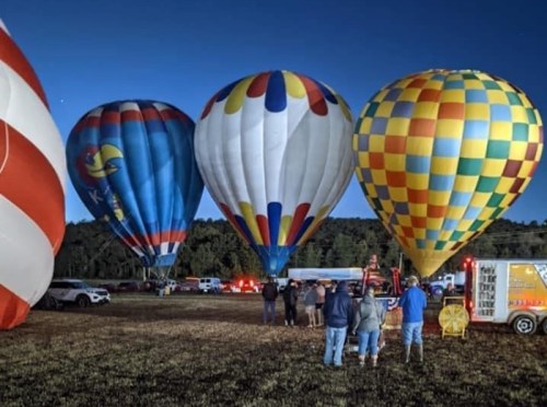 A group of colorful hot air balloons is illuminated at dusk, with people gathered nearby in a grassy field.