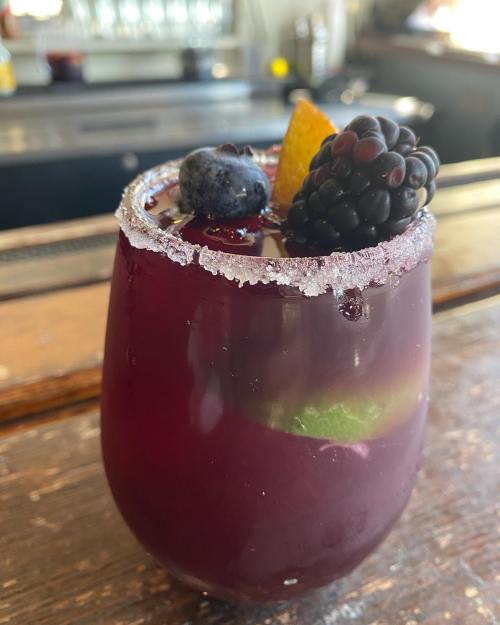 A colorful cocktail in a glass, garnished with blackberries, blueberries, and a slice of orange, rimmed with sugar.