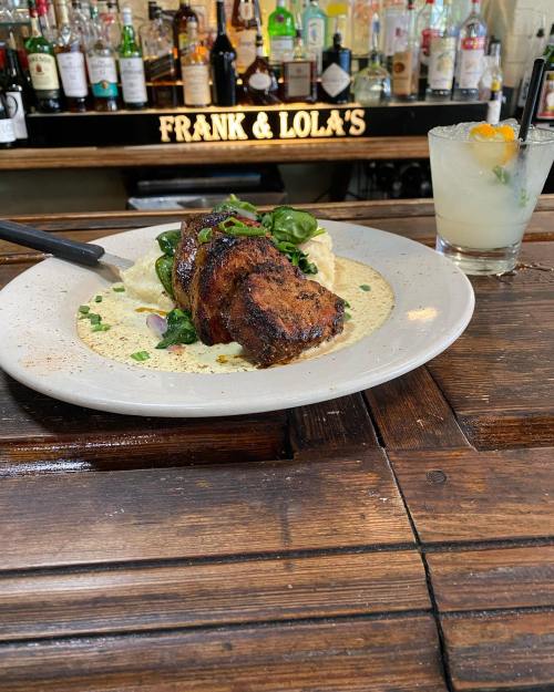 A plate of grilled meat with greens and sauce, set on a wooden table with a bar and cocktail in the background.