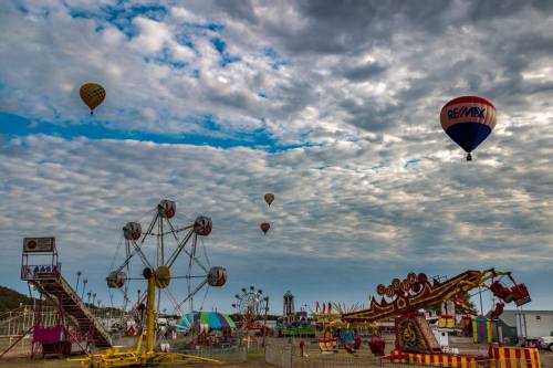A vibrant carnival scene with a Ferris wheel, rides, and colorful hot air balloons in a cloudy sky.