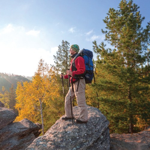 A hiker with a backpack stands on a rock, surrounded by trees and autumn foliage in a scenic outdoor landscape.