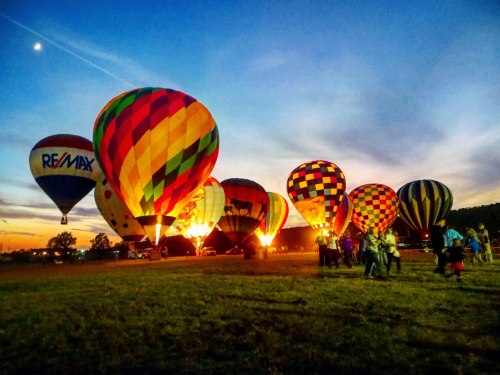 Colorful hot air balloons are illuminated at sunset, with people gathered on the ground in a grassy field.