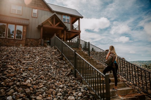 A woman climbs stone steps towards a large, rustic house under a cloudy sky.