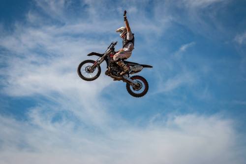 A motocross rider performs a jump, raising one hand in the air against a blue sky with wispy clouds.
