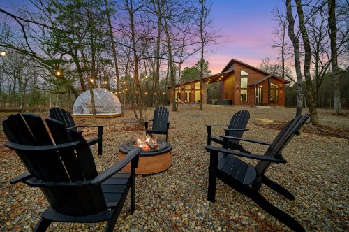 A cozy outdoor setting with black chairs around a fire pit, surrounded by trees and a modern cabin at sunset.