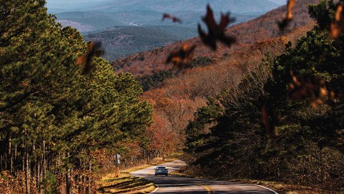 A winding road through autumn trees, with mountains in the background and a car driving along the scenic route.