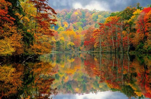 A serene lake surrounded by vibrant autumn foliage reflecting colorful trees and a cloudy sky.