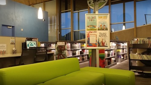 A cozy library interior featuring a green couch, bookshelves, and a poster for a children's art event.