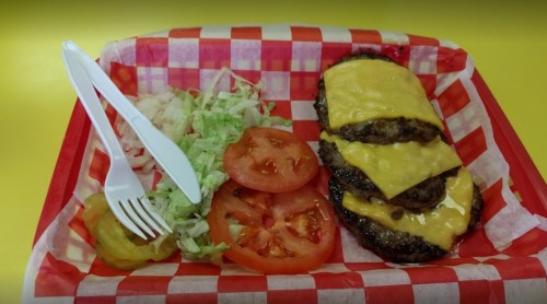 A tray with a cheeseburger featuring three patties, melted cheese, lettuce, and tomato slices on a red checkered background.