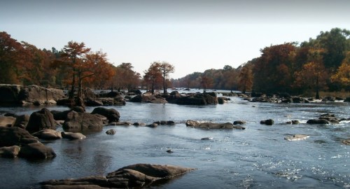 A serene river scene with rocky outcrops and autumn trees lining the banks under a clear blue sky.