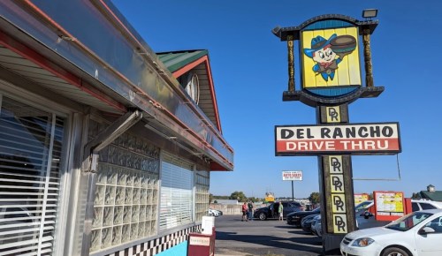 A colorful sign for Del Rancho Drive Thru next to a building with large windows and a red and white checkered pattern.