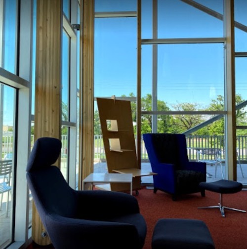 A bright, modern lounge area with large windows, featuring a blue chair, a black chair, and a wooden shelf.