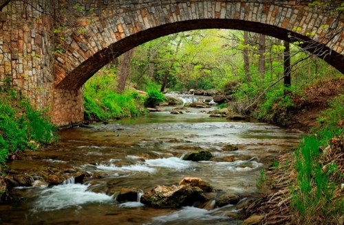 A serene stream flows under a stone arch bridge, surrounded by lush greenery and trees.