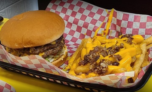 A juicy burger with toppings beside a plate of cheesy fries drizzled with sauce, all in a checkered basket.