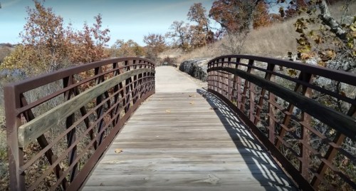 A wooden bridge with a metal railing spans a path through autumn foliage and trees.
