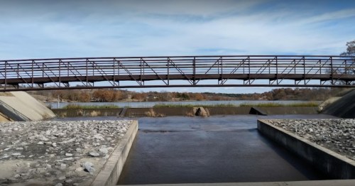 A metal bridge spans a calm river, surrounded by rocky banks and trees under a clear blue sky.