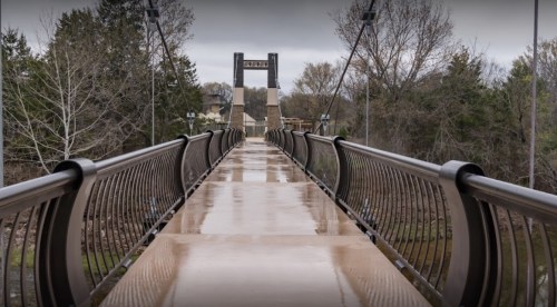 A wet, modern suspension bridge leads to a stone tower, surrounded by trees and a cloudy sky.