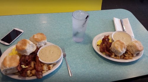 Two plates of breakfast with biscuits, bacon, potatoes, and gravy, alongside a glass of water and a smartphone.